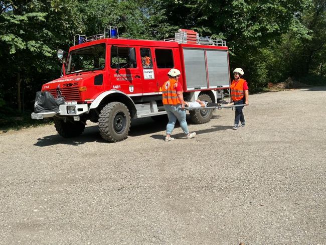 zu sehen ist ein Feuerwehrfahrzeug und zwei Jugendrotkreuzler mit einer Trage