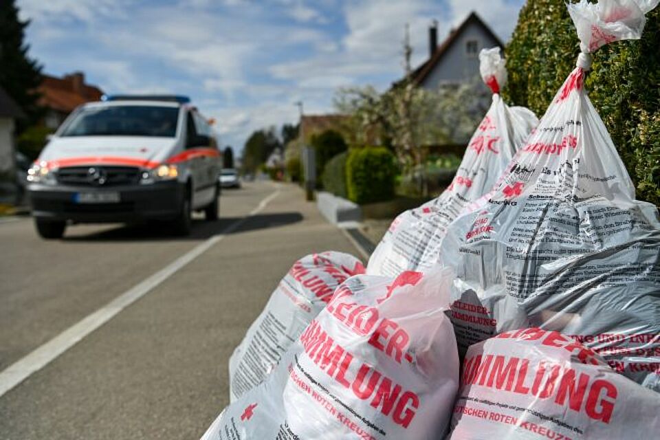 zu sehen ist ein DRK-Fahrzeug auf der Straße und ein Haufen mit Altkleidersäcken am Straßenrand bei einer Sammlung